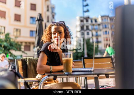 Frau aus dem Nahen Osten mit lockigen Haaren, die wegschaut, während sie tagsüber am Tisch mit Laptop und einem Glas Eiskaffee im Café im Freien sitzt Stockfoto