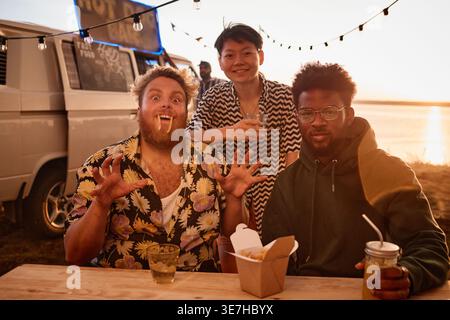 Gruppe von Freunden, die am Tisch mit Essen und Getränken sitzen und Spaß bei einer Strandparty haben Stockfoto