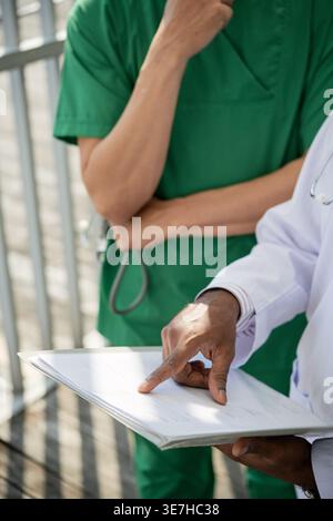 Zugeschnittenes Bild von asiatischen männlichen Ärzten im Scheueranzug und weißem Laborkittel, die auf das Dokument zeigen, während sie im Krankenhaus stehen Stockfoto