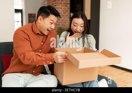 Überraschtes asiatisches Paar, das die Lieferbox öffnet, während es zu Hause auf dem Sofa im Wohnzimmer sitzt Stockfoto