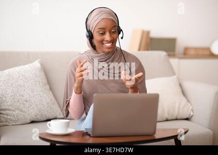 Schwarze muslimische Geschäftsfrau mit Headset, die Videoanrufe auf einem Laptop auf dem Sofa zu Hause führt Stockfoto