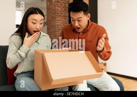 Überraschtes asiatisches Paar, das die Lieferbox öffnet, während es zu Hause auf dem Sofa im Wohnzimmer sitzt Stockfoto