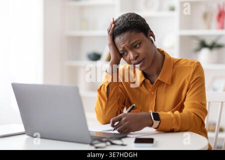 Gestresste schwarze Frau mit kurzen Haaren, die schnurlose Ohrhörer trägt, in Notebook schreibt und auf den Laptop-Bildschirm blickt, während sie mit Smartp am Tisch sitzt... Stockfoto