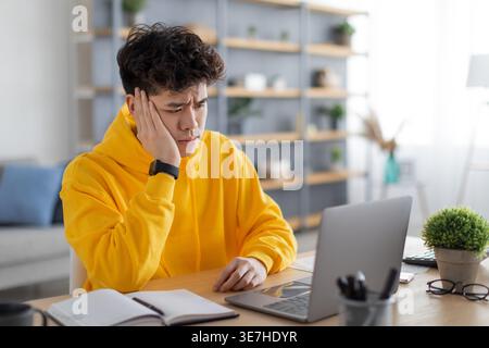Asiatischer Mann mit Hand auf Gesicht mit gelbem Hoodie und Blick auf Laptop, während er am Schreibtisch mit Notebook zu Hause sitzt Stockfoto