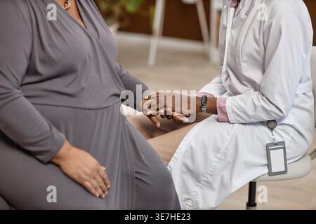 Zugeschnittenes Bild einer schwarzen Ärztin im Labormantel, die ihre Patientin mittleren Alters tröstet und Hand hält, während sie bei der Untersuchung zusammen sitzt... Stockfoto