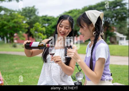 Thailändische Frau gießt Wein in Glas mit einer Freundin, die tagsüber auf einer Picknickdecke im Park sitzt Stockfoto