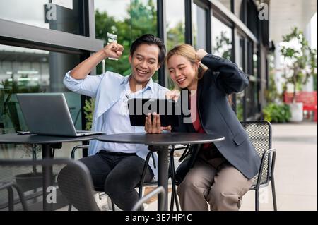 Thailändische Geschäftskollegen heben geschlossene Fäuste und blicken auf den Tablet-Bildschirm und feiern gute Nachrichten am Tisch im Café Stockfoto