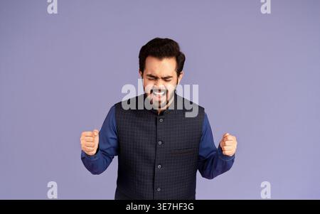 Indischer Mann mit geschlossenen Augen und geballter Faust, die vor violettem Hintergrund im Studio schreit Stockfoto