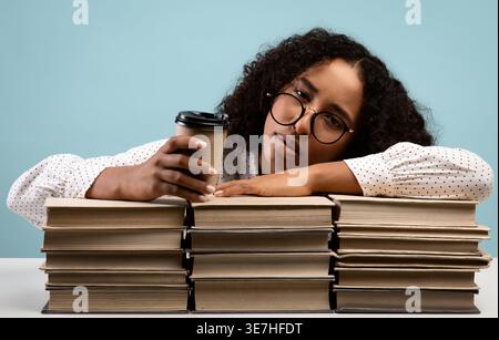 Müde Schwarzer weiblicher Teenager mit Brille, die eine Tasse Kaffee hält, lehnt sich auf Stapeln von Büchern, während er am Tisch vor blauem Hintergrund in s... Stockfoto