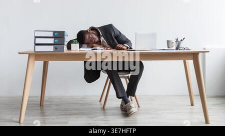 Müder Schwarzer Geschäftsmann mit Brille, der sich auf den Tisch stützt, mit Laptop, der vor der weißen Wand im Büro schläft Stockfoto