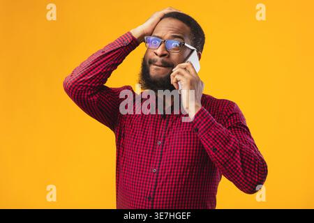 Verärgerter Schwarzer Mann mit Brille, der einen Anruf hat und wegschaut, während er vor gelbem Hintergrund im Studio steht Stockfoto