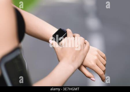 Zugeschnittenes Bild einer asiatischen Frau in Aktivkleidung, die die Zeit auf ihrer Uhr überprüft, während sie tagsüber auf der Straße steht Stockfoto
