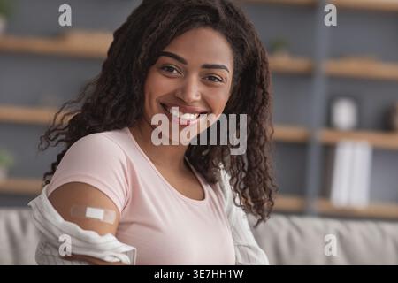 Lächelnde Schwarze Frau mit lockigem Haar, die Arm mit Pflaster im Wohnzimmer zu Hause zeigt Stockfoto