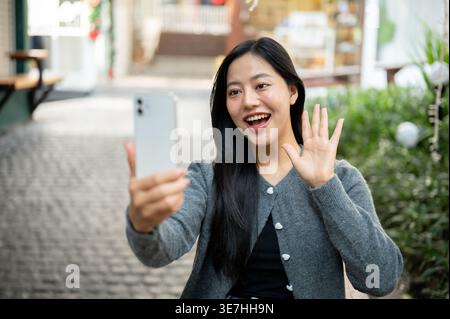 Fröhliche Thai-Frau mit Videoanruf auf dem Smartphone, die tagsüber im Freien sitzt Stockfoto