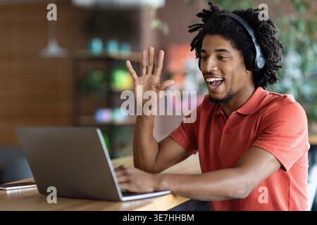 Schwarzer Mann mit Kopfhörern winkende Hand beim Videoanruf mit Laptop, der am Tisch im Café sitzt Stockfoto