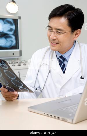 Chinesischer männlicher Arzt, der CT-Scan-Ergebnis hält, sitzt am Tisch im Krankenhaus Stockfoto