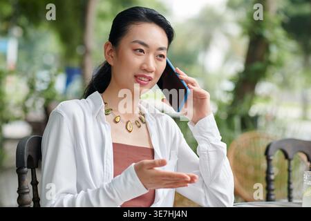 Asiatische Unternehmerin, die telefoniert und wegschaut, während sie im Café im Freien auf dem Stuhl sitzt Stockfoto