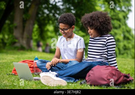 Schwarze Teenager beobachten auf dem Laptop, während sie im Park auf dem Gras sitzen Stockfoto