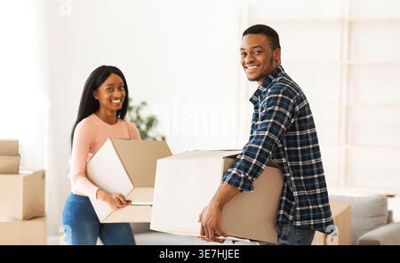 Ein lächelndes schwarzes Paar mit Pappschachteln, die sich zuhause im Wohnzimmer stehen Stockfoto