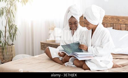 Lächelnde schwarze Frauen mit Handtüchern auf dem Kopf, die Bademäntel tragen, die Zeitschriften lesen, während sie zu Hause auf dem Bett sitzen Stockfoto