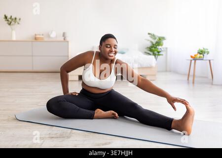Kurvige schwarze Frau, die Dehnübungen macht, während sie zu Hause im Schlafzimmer auf Yogamatte sitzt Stockfoto