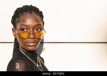 Schwarze Frau mit Zöpfen, die ein schwarzes Mesh-Oberteil und eine Sonnenbrille tragen und seitlich vor der weißen Wand wegschauen Stockfoto