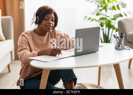 Schwarze Studentin mit Kopfhörern, die in einem Notebook schreiben und an einem Online-Kurs teilnehmen, während sie auf dem Boden neben dem Tisch sitzt und im Wohnzimmer um ... sitzt Stockfoto