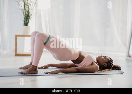 Schwarze Frau in Zöpfen, die Sportswear trägt, macht abs-Workout mit elastischem Band, während sie zu Hause auf einer Yogamatte liegt Stockfoto