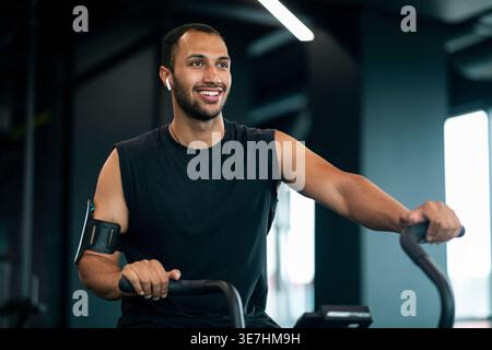 Schwarzer Mann mit Bart, der schnurlose Ohrhörer und Armband trägt und wegschaut, Training mit einem Crosstrainer im Fitnessstudio Stockfoto