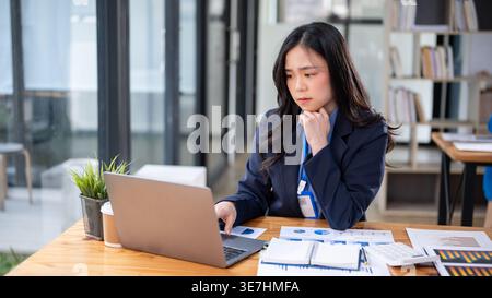 Besorgte asiatische Geschäftsfrau, die sich das Kinn zur Hand beugte und während der Arbeit im Büro am Schreibtisch mit Dokumenten auf ihren Laptop blickte Stockfoto
