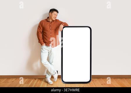 Asiatischer Mann, der Arm an einem großen Smartphone mit weißem Bildschirm lehnt, während er mit der Hand in der Tasche steht und die Beine zu Hause gegen die weiße Wand gekreuzt sind Stockfoto