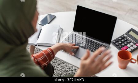 Zugeschnittenes Bild einer muslimischen Frau mittleren Alters im Hijab mit Laptop, während sie am Tisch mit Kaffeetasse, Taschenrechner, Smartphone und ... sitzt Stockfoto