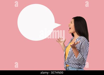 Asiatische Frau mit offenen Armen, die nach einer leeren Sprechblase schreien, während sie im Studio vor rosa Hintergrund steht Stockfoto