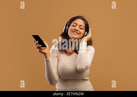 Verspielte arabische Dame mit Kopfhörern und Smartphone, die Musik hört und coole Playlist genießt, tanzt und sich über beigefarbenem Hintergrund zum Lieblingslied bewegt Stockfoto