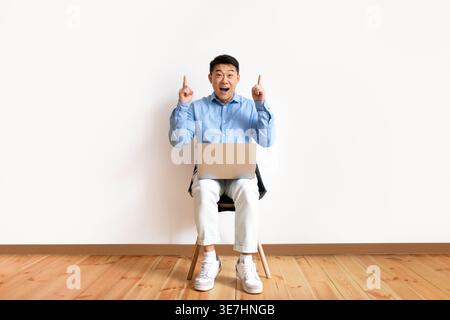 Asiatischer Mann, der die Finger nach oben zeigt, während er mit einem Laptop auf dem Stuhl an der weißen Wand zu Hause sitzt Stockfoto