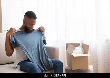 Schwarzer Mann, der T-Shirt vom Online-Kauf anprobiert, während er zu Hause auf dem Sofa sitzt Stockfoto