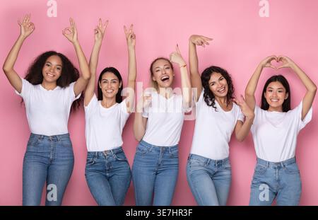 Verschiedene Frauengruppen in weißen Hemden und Jeans mit verschiedenen Handschildern, die im Studio vor rosafarbenem Hintergrund stehen Stockfoto