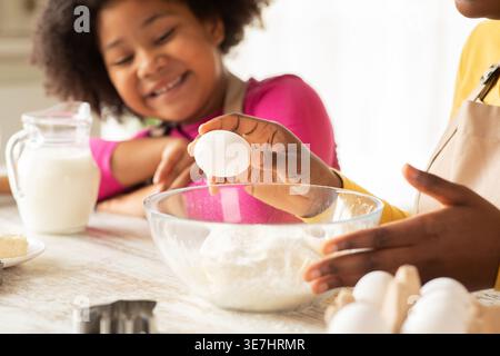Ein schwarzes Mädchen, das ihre Mutter ansieht, wie sie Ei in eine Schüssel knackte und Keksteig zubereitete, zusammen am Tisch in der Küche zu Hause sitzt Stockfoto