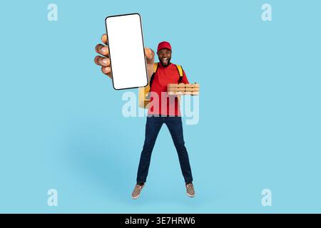 Schwarzer Liefermann mit offenem Mund, der eine Kappe trägt, die Thermotasche mit Pizzaschachteln hält und Smartphone mit leerem Bildschirm zeigt, während er springt... Stockfoto