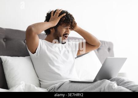 Gestresster bärtiger indischer Mann, der lockiges Haar berührt und Kopfschmerzen erlitt, während er mit Laptop auf dem Bett zu Hause im Schlafzimmer sitzt Stockfoto