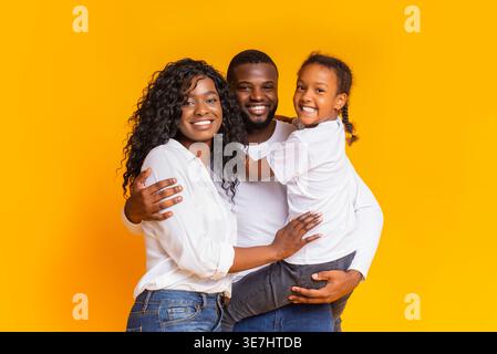 Die lächelnde Schwarze Familie umarmt sich gegenseitig mit dem Vater, der die Tochter trägt, steht vor gelbem Hintergrund im Atelier Stockfoto