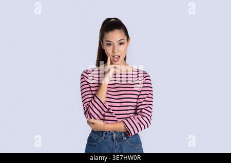Schwarze Frau mit offenem Mund in lässigem Outfit, die ihr Gesicht berührt, während sie im Studio vor grauem Hintergrund steht Stockfoto