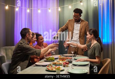 Schwarzer Mann im Business-Anzug, der Wein in Gläser gießt, der vor Freunden während der Dinnerparty drinnen steht Stockfoto