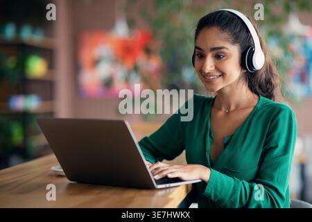 Indische Frau, die Kopfhörer trägt, indem sie einen Laptop benutzt, der am Tisch im Café sitzt Stockfoto