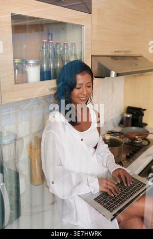 Fröhliche asiatische Frau, die auf der Küchenzeile sitzt und zu Hause am Laptop arbeitet Stockfoto