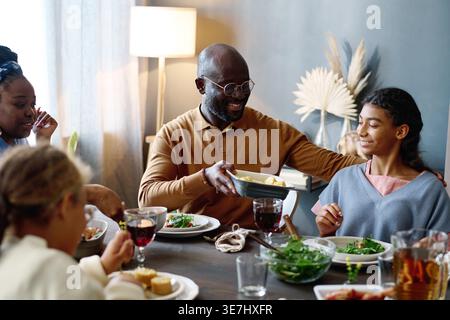 Schwarzer Vater in Brille, der Tablett mit gebackenem Mais an seine Tochter weitergibt, während er am Tisch sitzt und zu Hause ein Familienessen hat Stockfoto