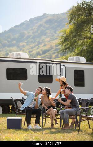 Lächelnde chinesische Freunde machen Selfie mit Bierflaschen, die auf Stühlen in der Nähe des Wohnwagens im Park sitzen Stockfoto