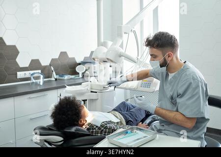 Schwarzer Junge, der mit kaukasischem männlichen Zahnarzt behandelt wird, während er auf einer Zahnarztklinik sitzt Stockfoto