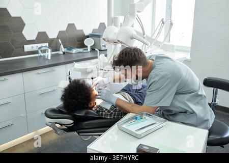 Schwarzer Junge, der mit kaukasischem männlichen Zahnarzt behandelt wird, während er auf einer Zahnarztklinik sitzt Stockfoto