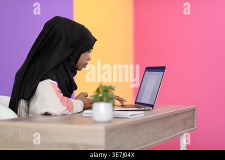 Ernsthafte schwarze muslimische Geschäftsfrau mit Nasenpiercing, die Hijab trägt und am Laptop arbeitet, während sie im Büro am Tisch sitzt Stockfoto